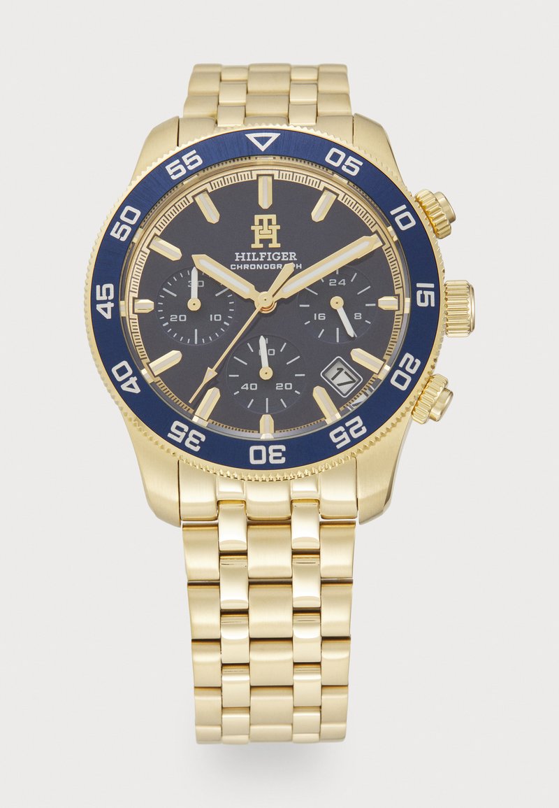 Auksinės spalvos chronografas su mėlynu bėgeliu, juodu ciferblatu ir trimis papildomais ciferblatais. Yra metalinė apyrankė ir datos langelis 4 valandos pozicijoje.