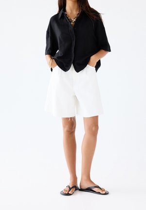 Chemise noire en lin à manches courtes avec boutons, associée à un short blanc taille haute et des tongs noires. Bijoux simples en accent.