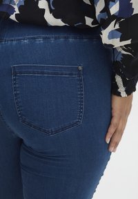Jeans de mezclilla azul oscuro con un solo bolsillo trasero, que presenta un diseño cosido y un pequeño botón de metal. Se aprecia una textura suave en la tela.