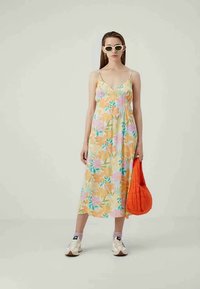 Floral maxi dress in pastel colors, featuring thin straps and a v-neck. Paired with an orange textured bag and white sneakers.