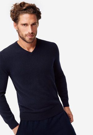 Marineblauer V-Ausschnitt-Pullover aus weichem Stoff, mit langen Ärmeln und tailliertem Design, kombiniert mit dunklen Hosen.