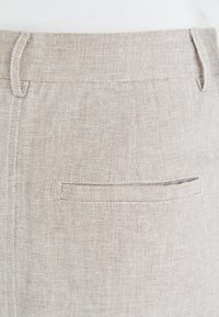 Nahaufnahme von beigen, strukturierten Hosen aus Stoff mit einer horizontalen Gesäßtasche mit Paspel und Gürtelschlaufen vor weißem Hintergrund.