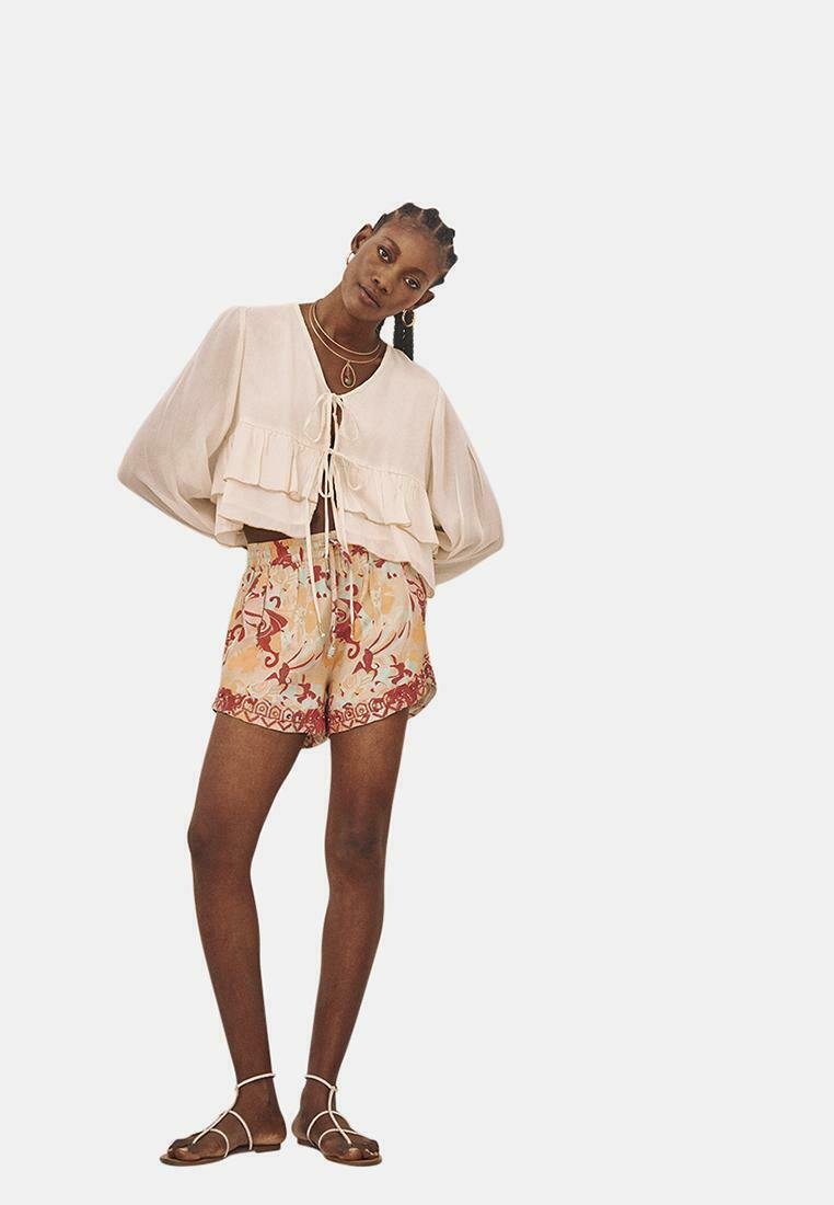 Top recortado de color beige claro con un dobladillo con volantes y un lazo en el frente, combinado con pantalones cortos de colores con un intrincado diseño floral.