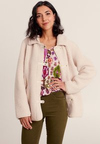 Giacca in pile color crema con colletto, dotata di chiusure a bottoni e tasche laterali. Indossata sopra una maglietta a fantasia floreale.