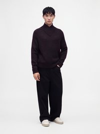 Homme debout portant un pull en maille bordeaux foncé, un pantalon noir à jambes larges et des baskets blanches sur un fond blanc uni.