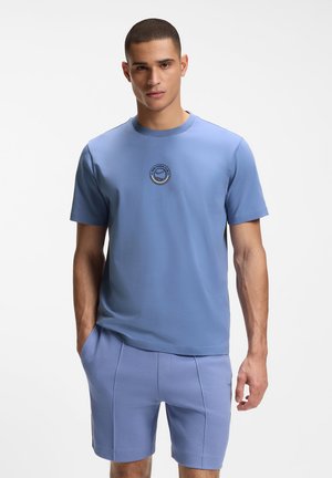 Mężczyzna ubrany w dopasowany jasnoniebieski tenisowy t-shirt i szorty, stojący z jedną ręką w kieszeni na jednolitym tle.