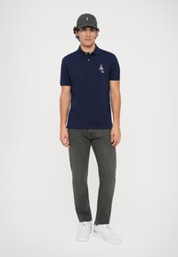 Polo azul marinho com mangas curtas e um pequeno design bordado no peito, combinado com calças cinza escuras e tênis brancos.