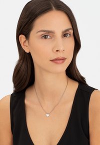 Zilveren ketting met een klein hartvormig hangertje, gedragen door een persoon met lang bruin haar, op een witte achtergrond. Gladde textuur, minimalistisch ontwerp.