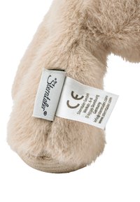 Fluffy, beige plush shoe with a textured surface. Features a light blue tag with care instructions and a CE mark. Round sole design.