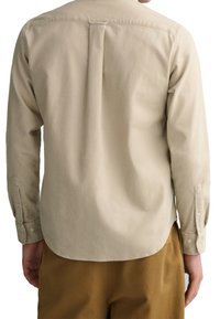 Långärmad skjorta, beige bomullstyg, detalj med veck bak, knäppta ärmslut, rundad nederkant, minimalistisk design. Buren med bruna byxor.