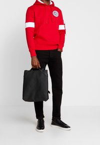 Červená mikina s bílými pruhy a logem, doplněná černými džínami a černými teniskami, držící černou tašku. Hladká textura látky.