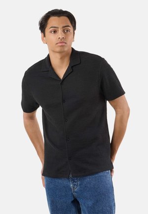 Man met kort zwart haar, gekleed in een zwart overhemd met korte mouwen en blauwe spijkerbroek, staand met de handen achter de rug.