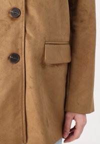 Manteau en daim marron avec des boutons foncés, devant boutonné, poche à rabat sur le côté, et une main visible près de l'ourlet.