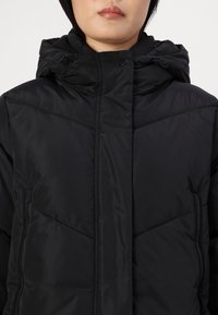 Giacca nera isolata con chiusura a zip, cappuccio e tasche laterali con zip. Presenta un motivo trapuntato e una texture liscia.