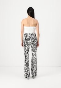 Femme aux longs cheveux noirs portant un haut blanc sans bretelles et un pantalon imprimé zèbre noir et blanc, debout face à un mur blanc.