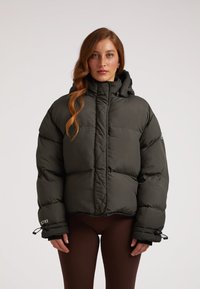 Giacca puffer verde scuro con colletto alto, chiusura con zip e polsini elastici. Dotata di cappuccio e lacci laterali per regolare la vestibilità.