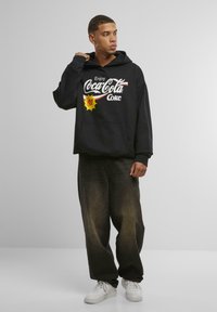Sort hoodie med "Coca-Cola" logo og grafik, frontlomme, parret med mørke, baggy jeans og hvide sneakers, sat op mod en ensfarvet baggrund.