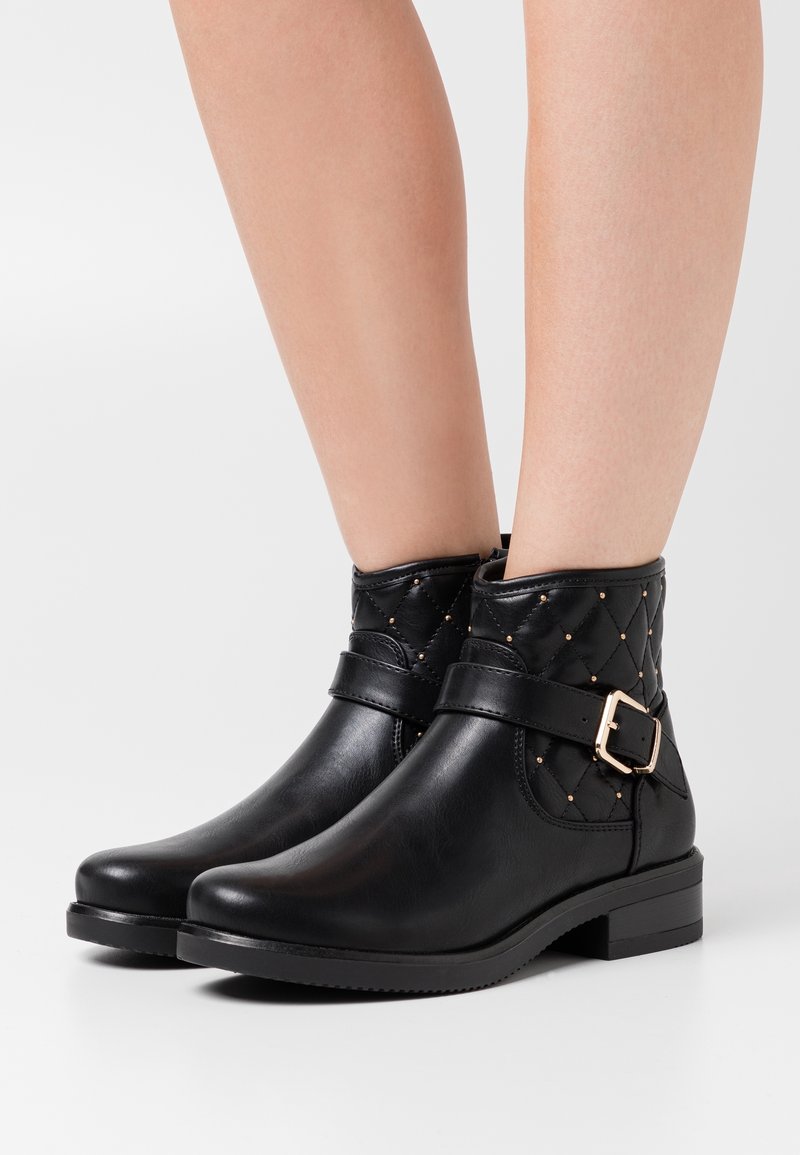 Bottes noires à chevilles avec tige matelassée matelassée, clous dorés, sangle latérale et boucle portées sur des jambes nues.
