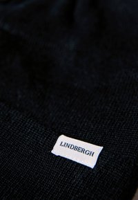 Tessuto nero con texture e un'etichetta bianca visibile che riporta la scritta "LINDBERGH" in caratteri neri. Il materiale appare morbido e tessuto.