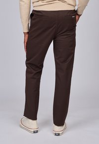 Pantalons en coton marron avec une coupe droite, dotés de deux poches arrière et de détails de couture subtils, associés à des chaussures blanc cassé.