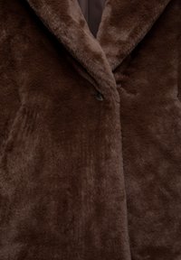 Rjava plašč iz umetnega krzna ima mehak, puhast teksturo, široke reverje in enojno zapiranje s gumbom. Oblikovanje poudarja toploto in udobje.