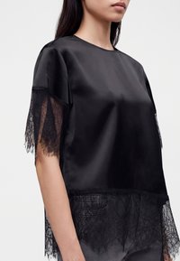 Femme portant un chemisier en satin noir à manches courtes avec des détails en dentelle noire transparente sur les manches et l’ourlet, sur un fond uni.