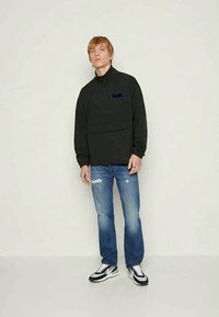 Un giovane che indossa un pullover nero con zip a metà, jeans blu strappati e sneakers bianco e nere, in piedi contro un muro chiaro e uniforme.