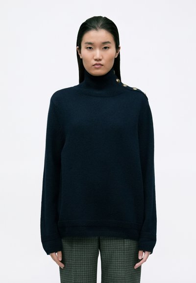 Pull à col roulé marine en matériau doux avec une coupe ajustée et des accents de boutons dorés à l'épaule, associé à un pantalon à carreaux.