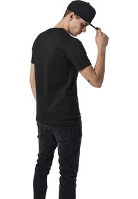 Camiseta de algodón negra de manga corta, con un diseño clásico de cuello redondo, combinada con jeans oscuros. El portador lleva una gorra negra inclinada hacia atrás.