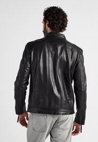 Schwarze Lederjacke mit glatter Textur, ausgestattet mit einem hohen Kragen und Reißverschlüssen an den Bündchen. Rückansicht, die die Nahtdetails hervorhebt.