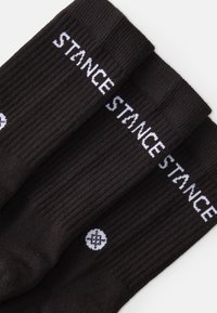 Svarta bomullssockor med ribbad textur, vitt "STANCE"-logotyp och en liten cirkulär vit grafisk detalj på sidan.