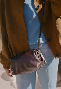 Borsa a tracolla in pelle marrone tenuta da una persona che indossa un maglione azzurro chiaro, pantaloni azzurri chiari e una giacca marrone con bottoni.