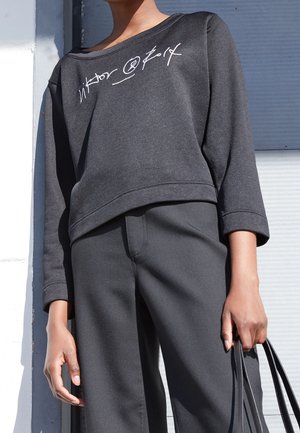 Osoba ubrana w ciemnoszarą krótką bluzę z białym napisem i dopasowane spodnie, trzymająca po bokach paski czarnej torebki.