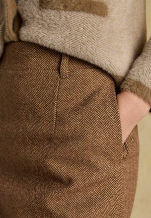 Detailný záber na ruku v vrecku, oblečenú v hnedých nohaviciach so vzorom rybej kosti a béžovom svetríku s textúrou rukáva.