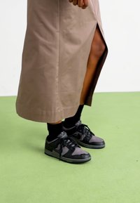 Fekete és szürke sneaker cipők velúr textúrával; fekete zoknikkal viselve, és egy oldalsó hasítékos, homokszínű, boka hosszig érő szoknyával. Zöld padló.