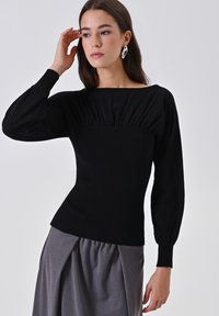 Donna con lunghi capelli scuri, che indossa un top nero a maniche lunghe arricciato e una gonna plissettata grigia, mentre sistema i capelli vicino al volto, su uno sfondo neutro.