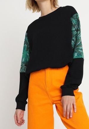 Kvinde iført sort langærmet top med grønne bladmønstre på ærmerne og lyse orange højtaljede bukser mod en ensfarvet baggrund.