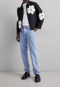 Chaqueta negra con estampado floral de flores blancas, llevada sobre una camiseta blanca lisa. Los jeans azules y los zapatos negros completan el conjunto, con un ajuste relajado.