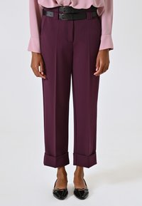 Pantalons ajustés violets avec des ourlets repliés, associés à une ceinture noire à double boucle et un chemisier rose pâle. Texture de tissu lisse.