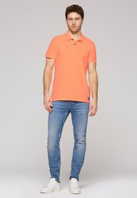 Camisa polo de color naranja brillante con un pequeño bolsillo en el pecho, combinada con jeans de mezclilla azul claro y zapatillas blancas. Mangas cortas y color sólido.