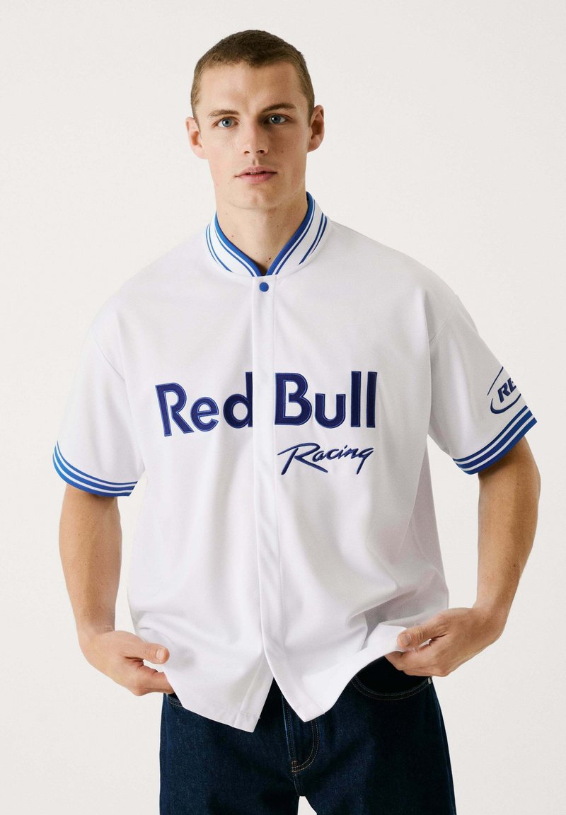 Witte baseball-stijl shirt met blauwe accenten. Bevat het "Red Bull Racing" logo en knopen, korte mouwen met gestreepte manchetten, gladde textuur.