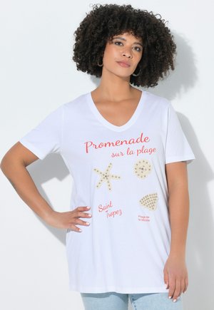 Kvinde med krøllet hår iført en hvid V-hals T-shirt med skaller og søstjerne design og rød fransk tekst, der refererer til Saint Tropez strand.