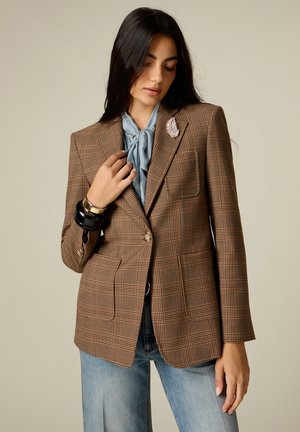 Mujer que lleva una blazer marrón de cuadros con una blusa con pañuelo azul claro, vaqueros azules claros, pulseras negras y doradas, y un broche decorativo en la solapa.