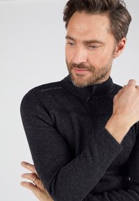 Sudadera de cremallera negra con cuello alto, hecha de un material texturizado. Presenta detalles cosidos en los hombros y un logo en el lateral.