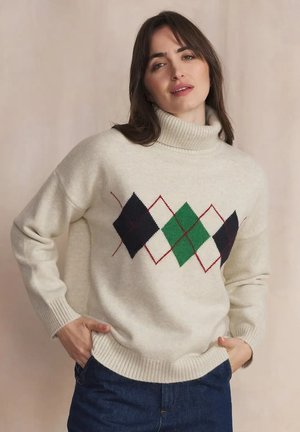 Femme portant un pull à col roulé crème avec un motif losanges noir, vert et blanc, mains dans les poches, sur fond beige uni.