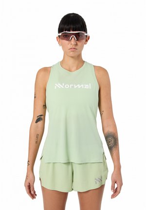 Persona che indossa una canotta verde chiaro senza maniche con la scritta "Normale" e pantaloncini abbinati, con occhiali da sole e tatuaggi visibili sulle braccia, rivolta verso il davanti.