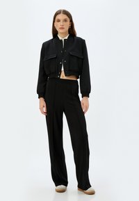 Chaqueta bomber negra recortada con dos bolsillos frontales y puños elásticos, combinada con pantalones de pierna ancha negros de talle alto y zapatos claros.