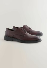 Chaussures en cuir bordeaux foncé avec une surface texturée, bout rond et un design à laçage à cinq œillets ; semelle en caoutchouc noire ajoutant un contraste.