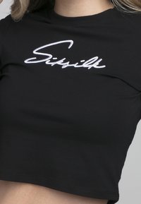Black cropped T-shirt featuring white embroidered "Siksilk" logo. Soft fabric with a smooth texture, short sleeves, and a fitted design.