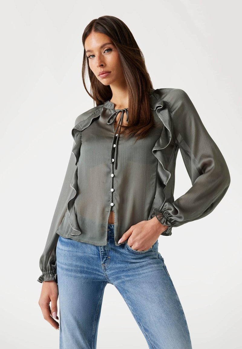 Bluză gri transparentă cu accente volanate, mâneci lungi, detaliu cu fundă la gât și butoane în față, asortată cu blugi pentru un look casual.